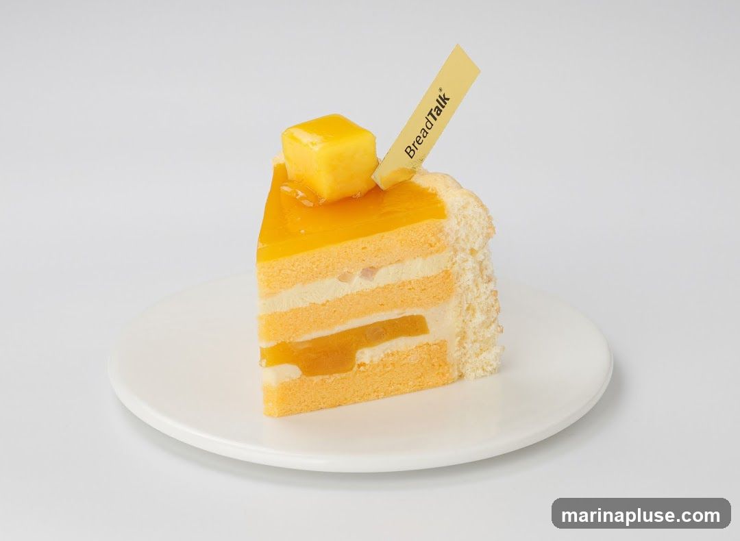 A slice of layered mango cake with orange sponge cake, white cream, and mango filling, topped with a mango cube and a yellow flag with "BreadTalk" written on it. The cake is served on a white plate against a white background.