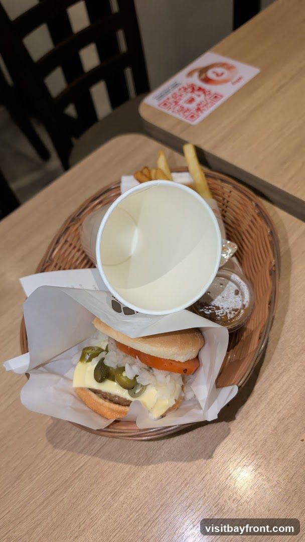 A close-up shot of a burger, fries, and a cola drink on a wooden table. The burger is open-faced, showing a thick patty with lettuce and sauce. The fries are in a white paper wrapper, and the cola is in a clear glass with ice. The background is a textured gray wall.