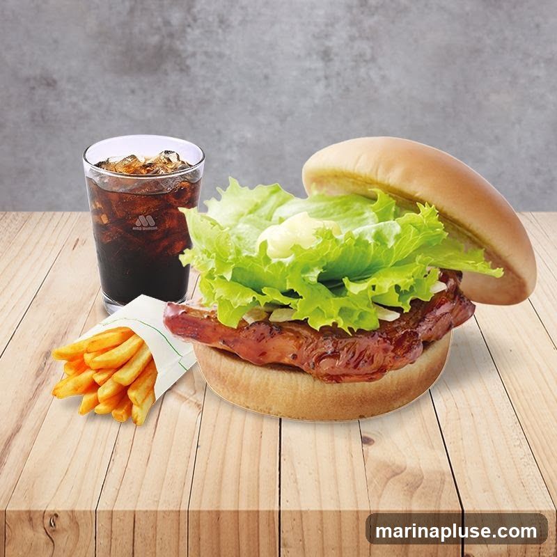 A close-up shot of a burger, fries, and a cola drink on a wooden table. The burger is open-faced, showing a thick patty with lettuce and sauce. The fries are in a white paper wrapper, and the cola is in a clear glass with ice. The background is a textured gray wall.