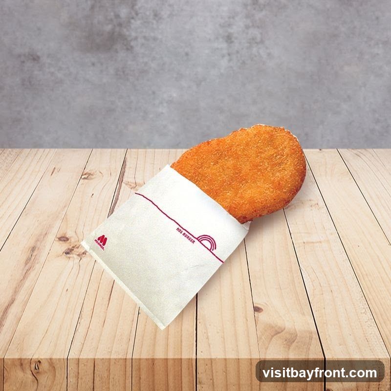 A golden-brown, oval-shaped fried patty is partially wrapped in a white paper sleeve. The sleeve has a red outline and a small red logo on the left side. Text on the sleeve reads "NOS BURGER" and features a stylized rainbow graphic. The patty and wrapper rest on a light-colored wooden surface, with a textured gray background behind it.