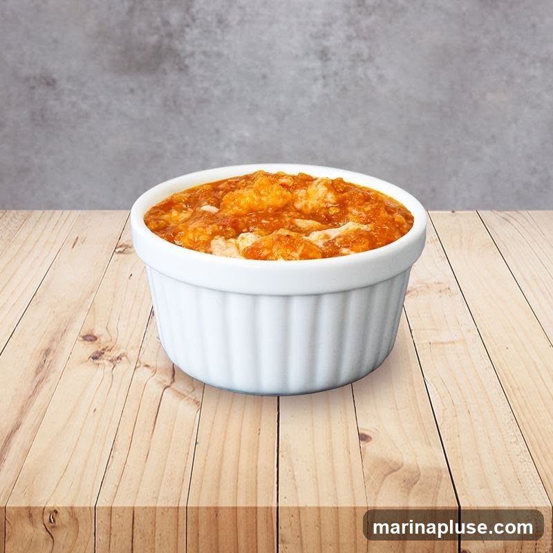 A white ramekin filled with a chunky orange-colored food, possibly a stew or casserole, sits on a light-colored wooden table. The background is a textured gray wall.