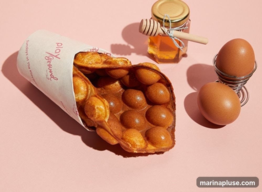 A golden-brown bubble waffle is presented in a paper cone with the text "Playground" visible. Beside it, a small jar of honey with a wooden dipper sits next to two brown eggs, one in an egg cup. The background is a soft pink.