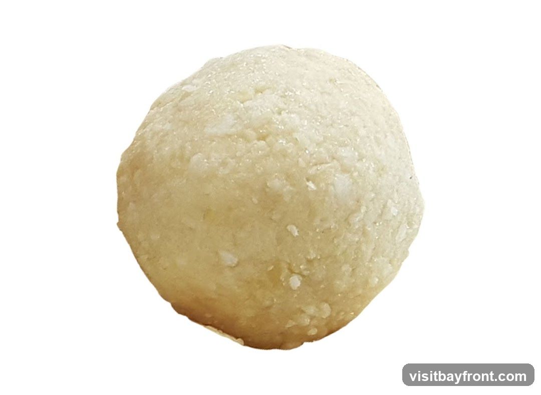 A close-up, isolated shot of a single, round, light-colored sweet, likely a laddu. The surface appears slightly textured with small, granular particles, possibly sugar or desiccated coconut. The sweet is a pale yellow or off-white color and is presented against a plain white background.