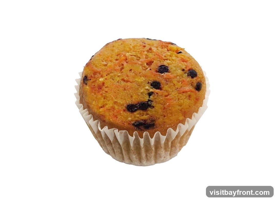 A close-up, isolated shot of a carrot muffin in a white paper liner. The muffin has a golden-brown, textured top with visible flecks of orange carrot and dark chocolate chips.