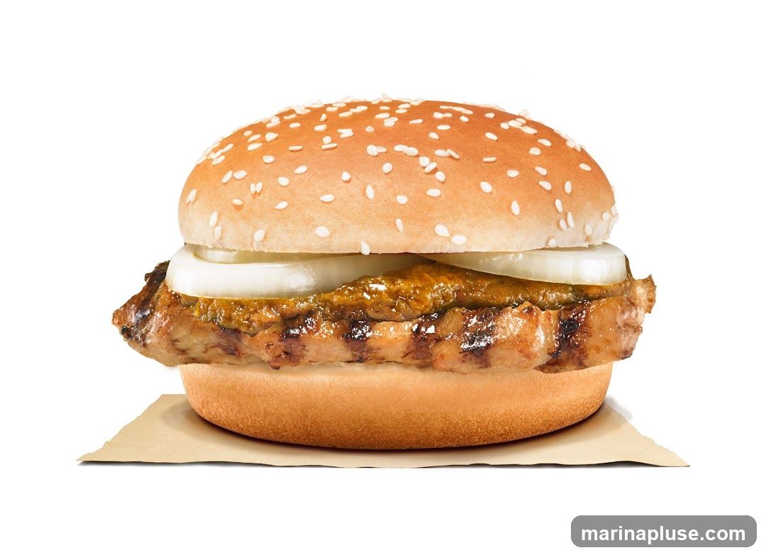 A close-up studio shot of a grilled chicken burger on a white background. The burger has a sesame seed bun, grilled chicken patty, onions, and a sauce. It is served on a piece of brown paper.