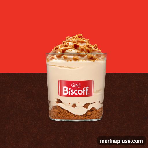 A clear glass filled with a layered dessert. The bottom layer appears to be crushed Biscoff cookies, followed by a creamy white filling, and topped with more cream, a drizzle of caramel sauce, and crumbled Biscoff cookies. A red label with the Lotus Biscoff logo is affixed to the glass. The background is split horizontally, with a red section at the top and a dark brown textured section at the bottom.