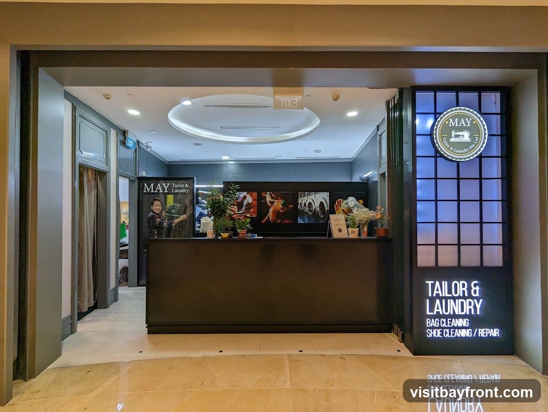 The exterior of a tailor and laundry shop. A dark counter is visible with some decorative items and framed pictures. To the right, a vertical sign displays the shop's name and services: TAILOR & LAUNDRY, BAG CLEANING, SHOE CLEANING / REPAIR. A circular logo with a sewing machine is above the name.