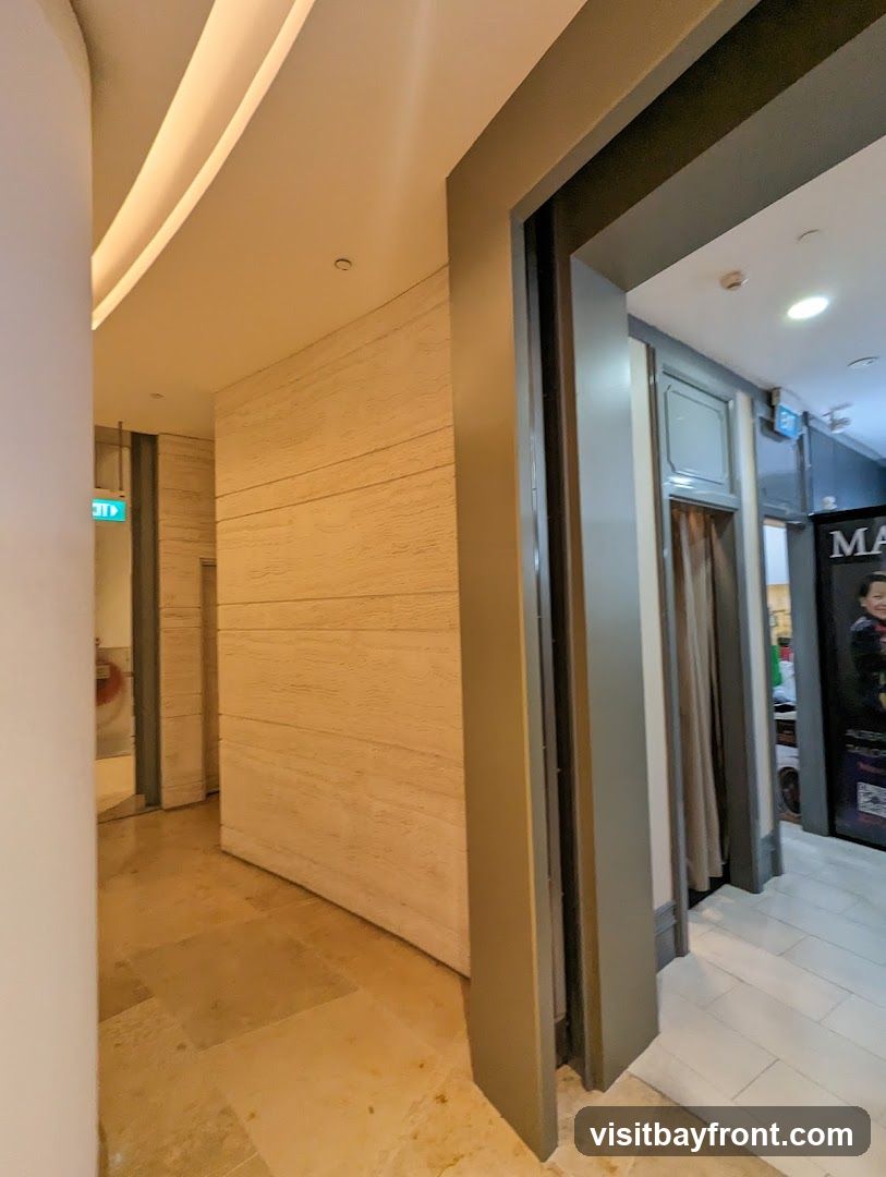 A view down a well-lit interior hallway with a textured wall on the left and an open doorway on the right. An illuminated exit sign is visible in the distance. The doorway on the right leads to a space with a curtained area and a poster that partially shows the text "MA" and "TAILOR".
