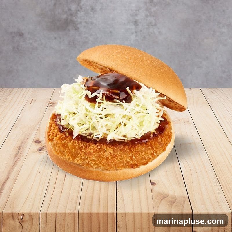 A close-up shot of a katsu burger on a wooden table. The burger has a golden-brown, crispy katsu patty, topped with shredded cabbage and drizzled with a dark sauce. The top bun is slightly ajar, revealing the fillings. The background is a textured gray wall.