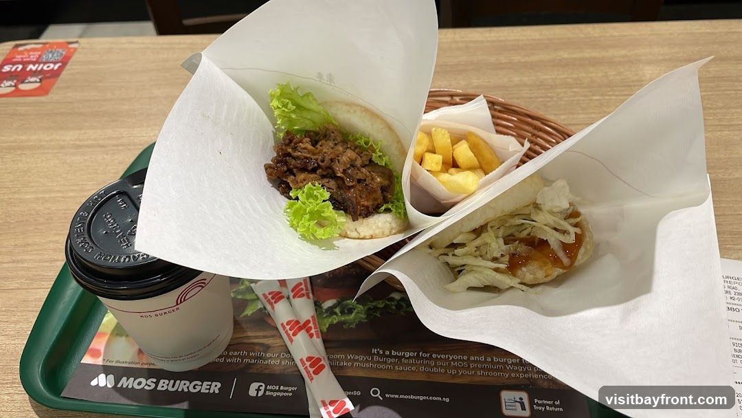A white container filled with french fries and onion rings is shown. Two glasses of Coca-Cola, labeled "Original Taste Less Sugar," are placed in front of the container. The Mos Burger logo is visible in the upper right corner.