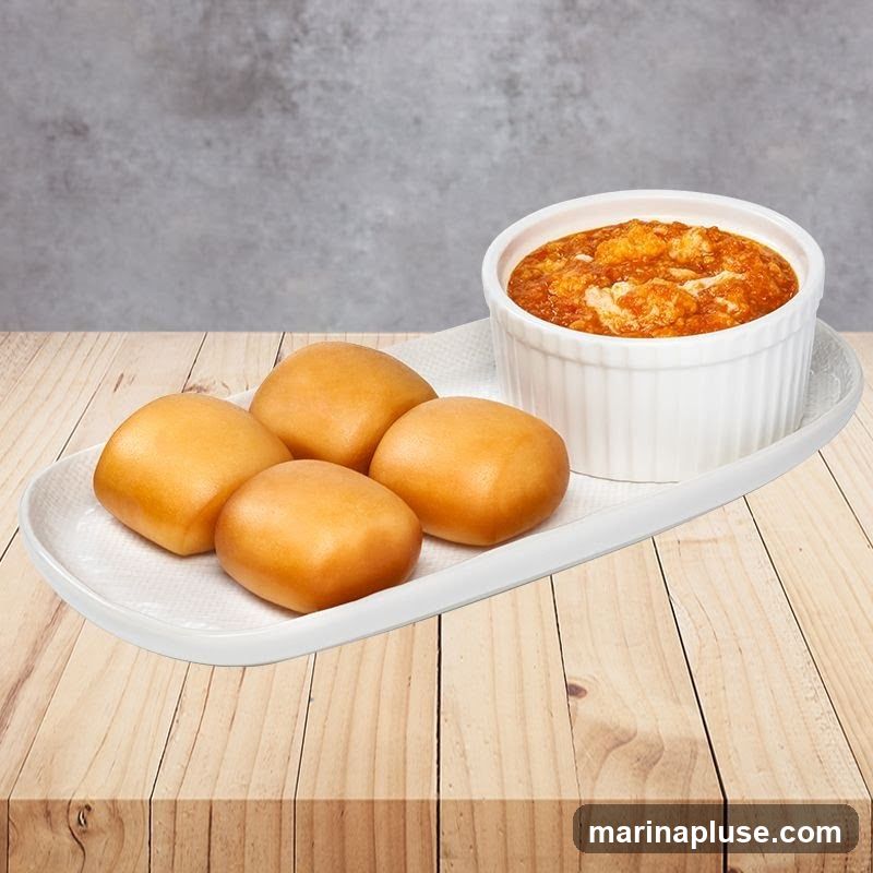 Four golden-brown fried mantou buns are arranged on a white rectangular plate with a white napkin. To the right of the buns, a white ramekin holds a savory dip with visible chunks of food, possibly meat or seafood, in a reddish-orange sauce. The plate and ramekin are set on a light-colored wooden surface, with a textured gray background behind.