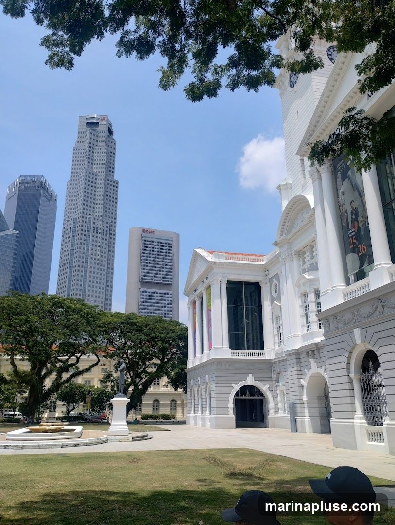 A view of the Victoria Theatre and Concert Hall in Singapore, with modern skyscrapers in the background. The white colonial-era building features classical architecture with columns and arches. A statue stands in a grassy area in front of the building. The sky is clear blue with some clouds.