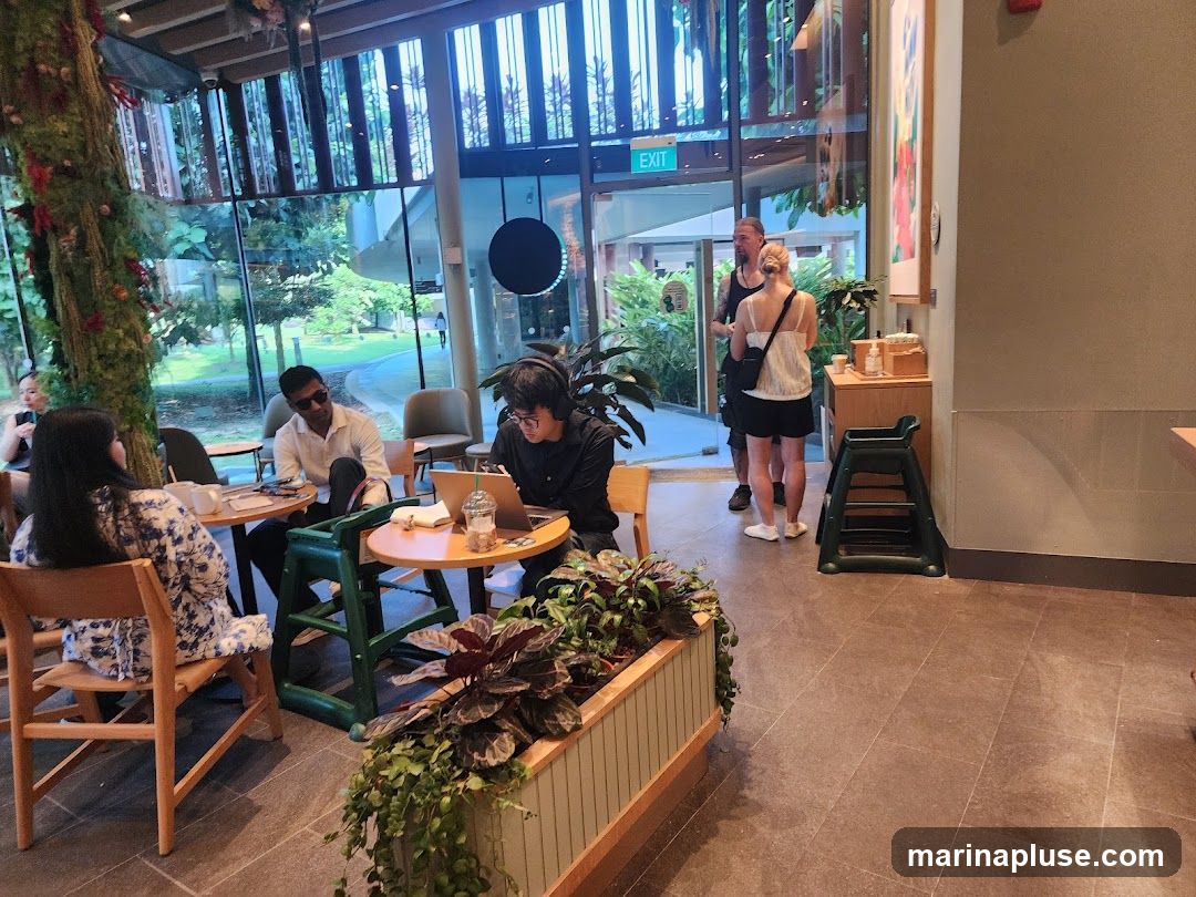A group of people are seated and standing inside a cafe with large windows overlooking a green outdoor area. One person is working on a laptop, while others are conversing. The cafe has a modern design with plants and artwork visible.