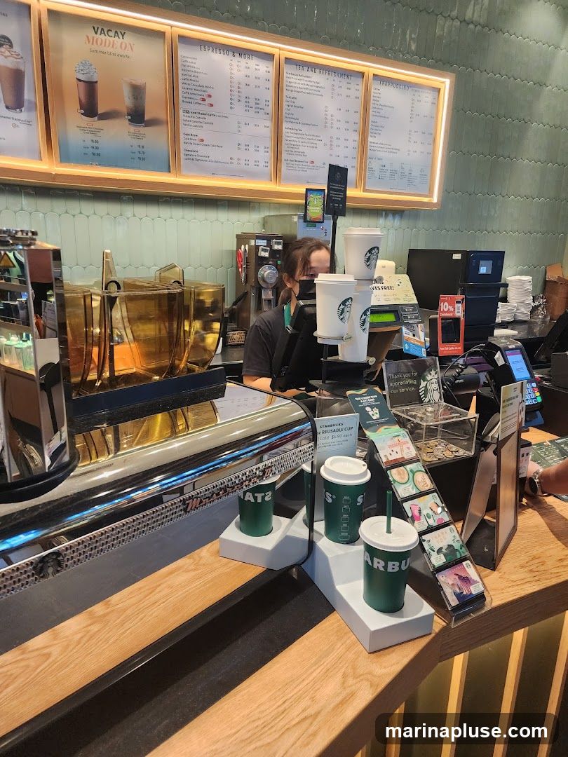 A Starbucks counter with a barista wearing a mask behind it. There are menu boards above, displaying various drinks and promotions. On the counter, there are reusable cups, a card display, and a cash register area.