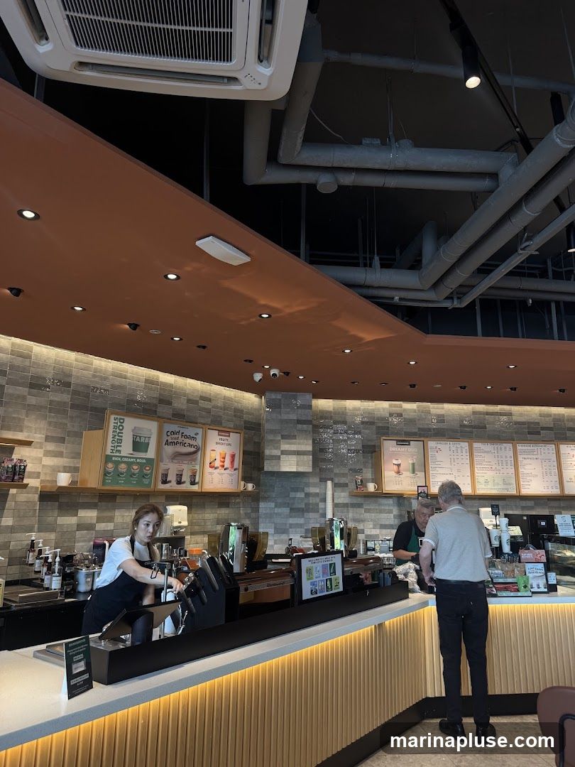 A view of a coffee shop counter with baristas preparing drinks and customers ordering. There are menus displayed on the wall behind the counter, showing various coffee and tea options. The interior features modern decor with tiled walls and wooden accents.