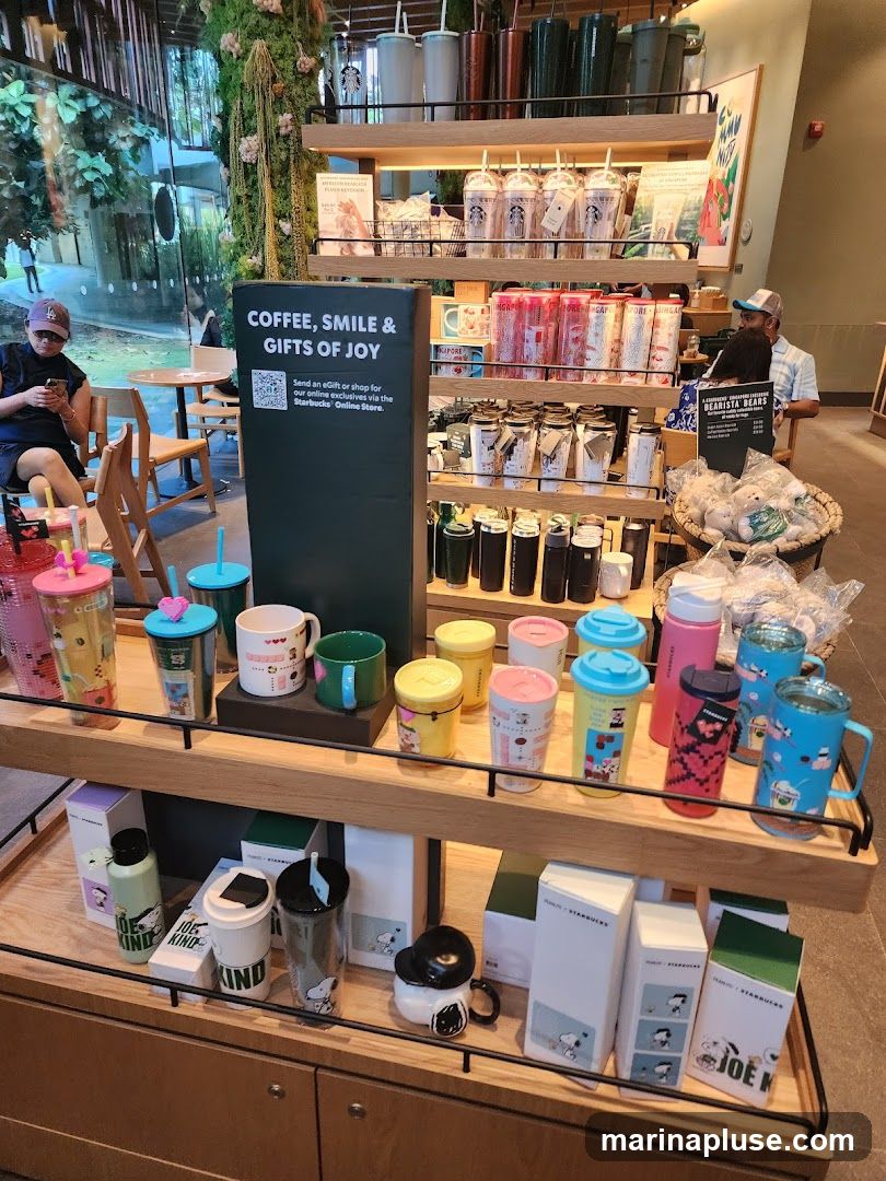 A display of Starbucks merchandise including tumblers, mugs, and other drinkware. A sign reads "COFFEE, SMILE & GIFTS OF JOY" and directs customers to send an eGift or shop online. Several customers are visible in the background, one sitting and looking at their phone, and another standing near a display of plush bears.