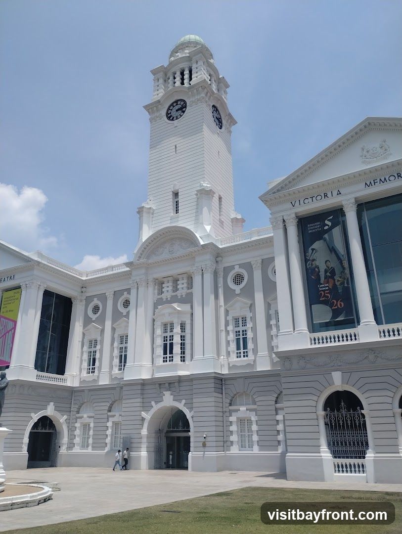 A grand white building with a prominent clock tower and classical architectural details. The facade features arched entrances, large windows, and decorative moldings. A banner for the Singapore Symphony Orchestra's concert season is visible on one side.