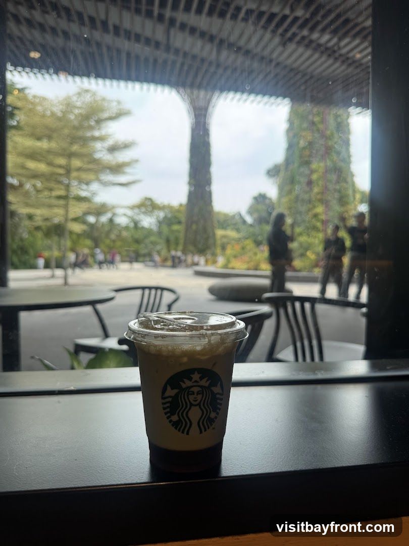 A Starbucks cup with the logo is placed on a dark surface, with a view of the Gardens by the Bay in Singapore through a window. The Supertrees and surrounding greenery are visible in the background, along with some people walking around.
