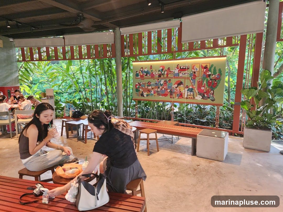A group of people are seated at tables in a cafe, some enjoying drinks. The cafe has an open-air feel with large windows or openings looking out onto dense green foliage. A colorful mural depicting various figures is displayed on one wall.