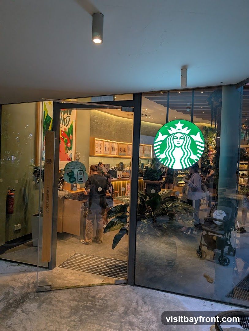 The exterior of a Starbucks coffee shop. A large green Starbucks logo is visible on the glass door. Inside, customers and staff are visible, along with a counter and display cases. Plants are placed near the entrance.
