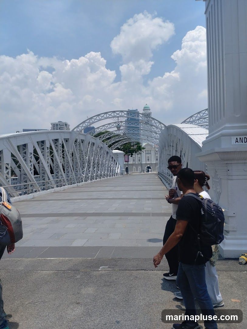 A group of people are walking across a white metal bridge on a sunny day. The bridge has an arched structure and is lined with decorative railings. In the background, there are buildings and a cloudy blue sky. One person in the foreground is wearing a black t-shirt and jeans, carrying a backpack. Another person is wearing a grey backpack.