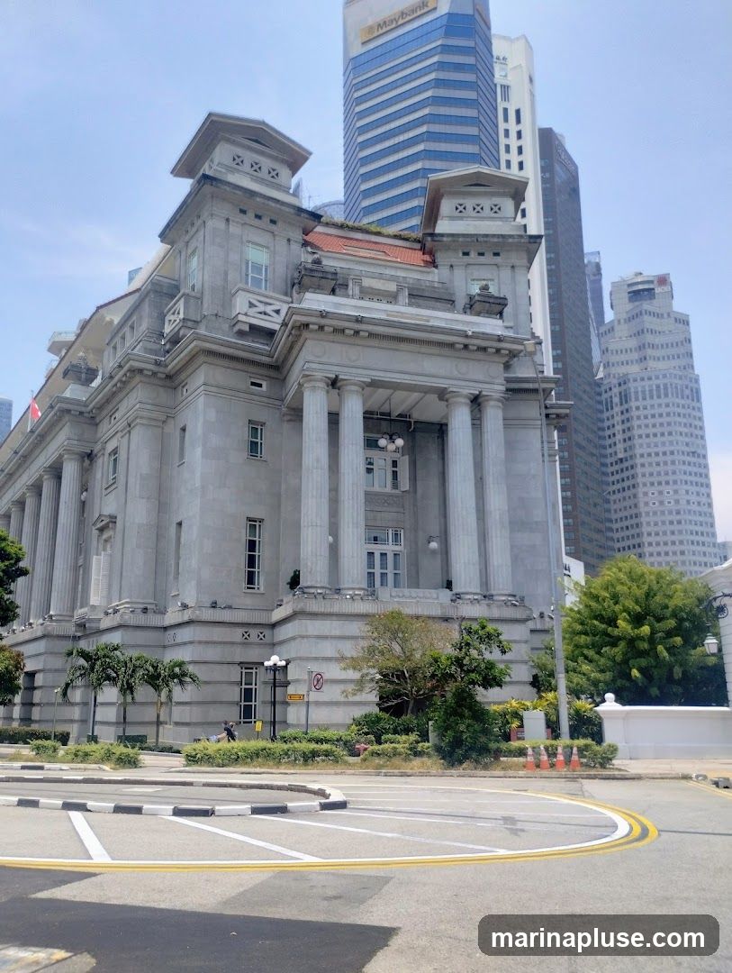 A grand, classical stone building with columns and ornate detailing stands prominently in the foreground. Behind it, modern skyscrapers pierce the sky, including a building with a "Maybank" logo. The scene is set on a sunny day with a clear blue sky. The foreground shows a street with road markings and some greenery.
