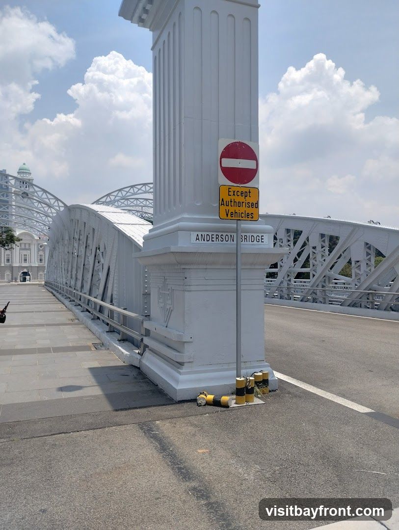 A view of the Anderson Bridge on a sunny day with a clear blue sky and scattered clouds. The bridge is white with intricate metalwork. A large white pillar stands in the foreground, adorned with a "No Entry" sign and a yellow sign stating "Except Authorised Vehicles". The name "ANDERSON BRIDGE" is inscribed on the base of the pillar. The road surface and a pedestrian walkway are visible.