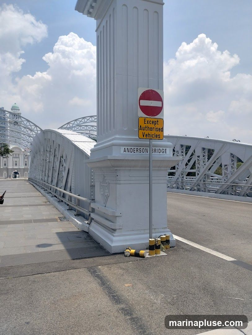 A view of the Anderson Bridge on a sunny day with a clear blue sky and scattered clouds. The bridge is white with intricate metalwork. A large white pillar stands in the foreground, adorned with a "No Entry" sign and a yellow sign stating "Except Authorised Vehicles". The name "ANDERSON BRIDGE" is inscribed on the base of the pillar. The road surface and a pedestrian walkway are visible.