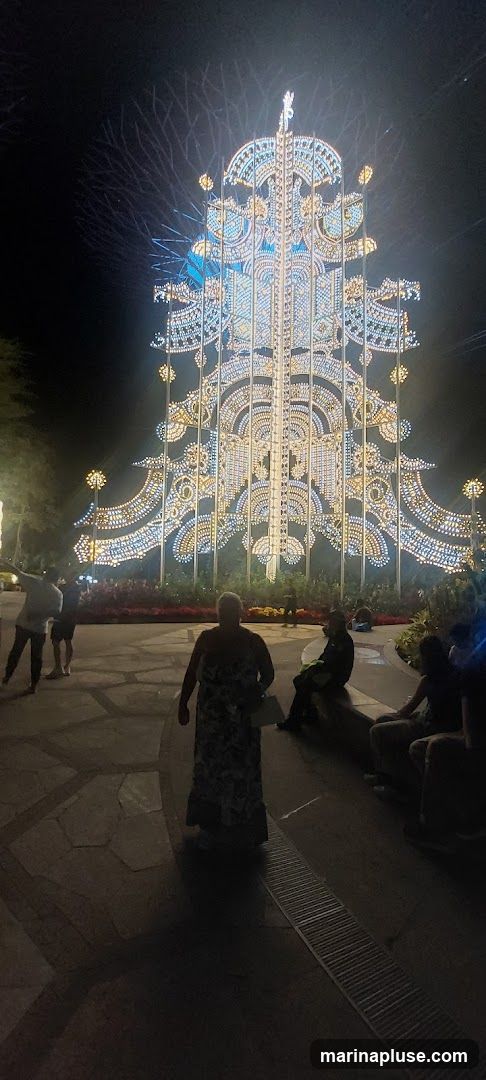 A large, ornate Christmas tree structure is illuminated with thousands of white and blue lights at night. The tree has intricate patterns and decorative elements. Several people are visible in the foreground and midground, some walking and others sitting on benches, observing the display. The ground is paved with stone tiles.