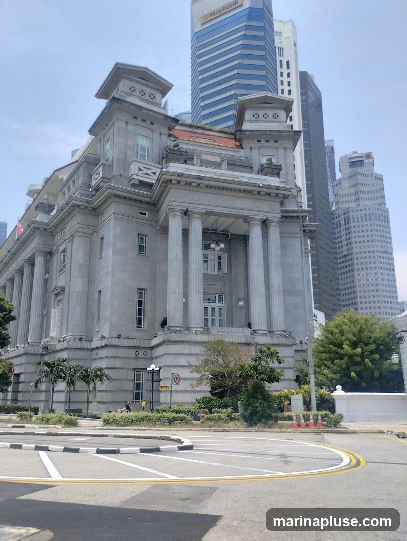A grand, classical-style building with columns and ornate details stands prominently in the foreground. Behind it, modern skyscrapers rise into the sky, including one with a "Maybank" sign. The scene is set on a clear day with a road and traffic cones visible in the lower portion of the image.