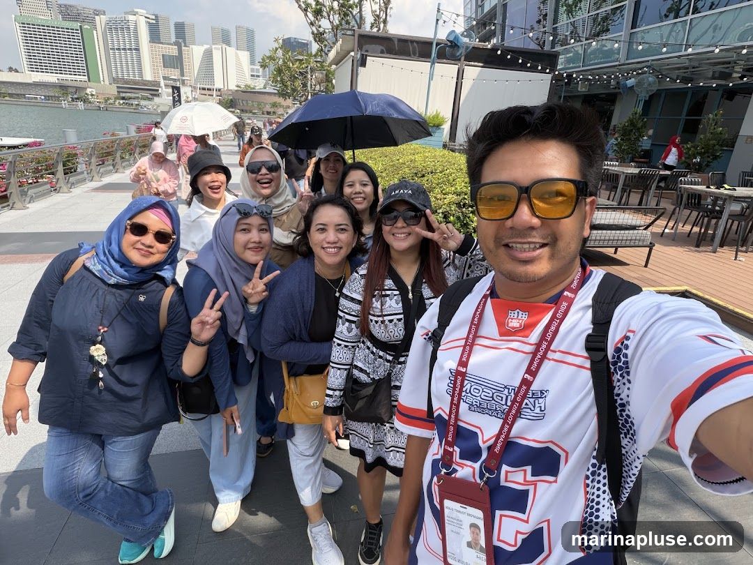 A group of nine people, mostly women, are posing for a selfie outdoors on a sunny day. They are standing on a paved walkway with a body of water and city skyline in the background. Some people are holding umbrellas. The person in the foreground on the right is wearing a white jersey with red and blue accents and yellow sunglasses. The others are dressed in casual attire. The background shows modern buildings and an outdoor seating area with tables and chairs.