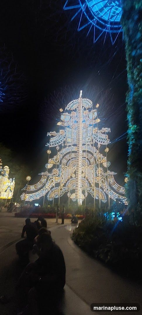 A large, intricately decorated Christmas tree made of lights stands tall against a dark night sky. The tree is adorned with numerous small lights in yellow and blue, creating a dazzling display. In the foreground, several people are seated or standing, observing the spectacle. To the right, a large, dark, leafy structure partially obscures the view. In the background, other illuminated structures and trees can be seen, suggesting a festive outdoor event or park.
