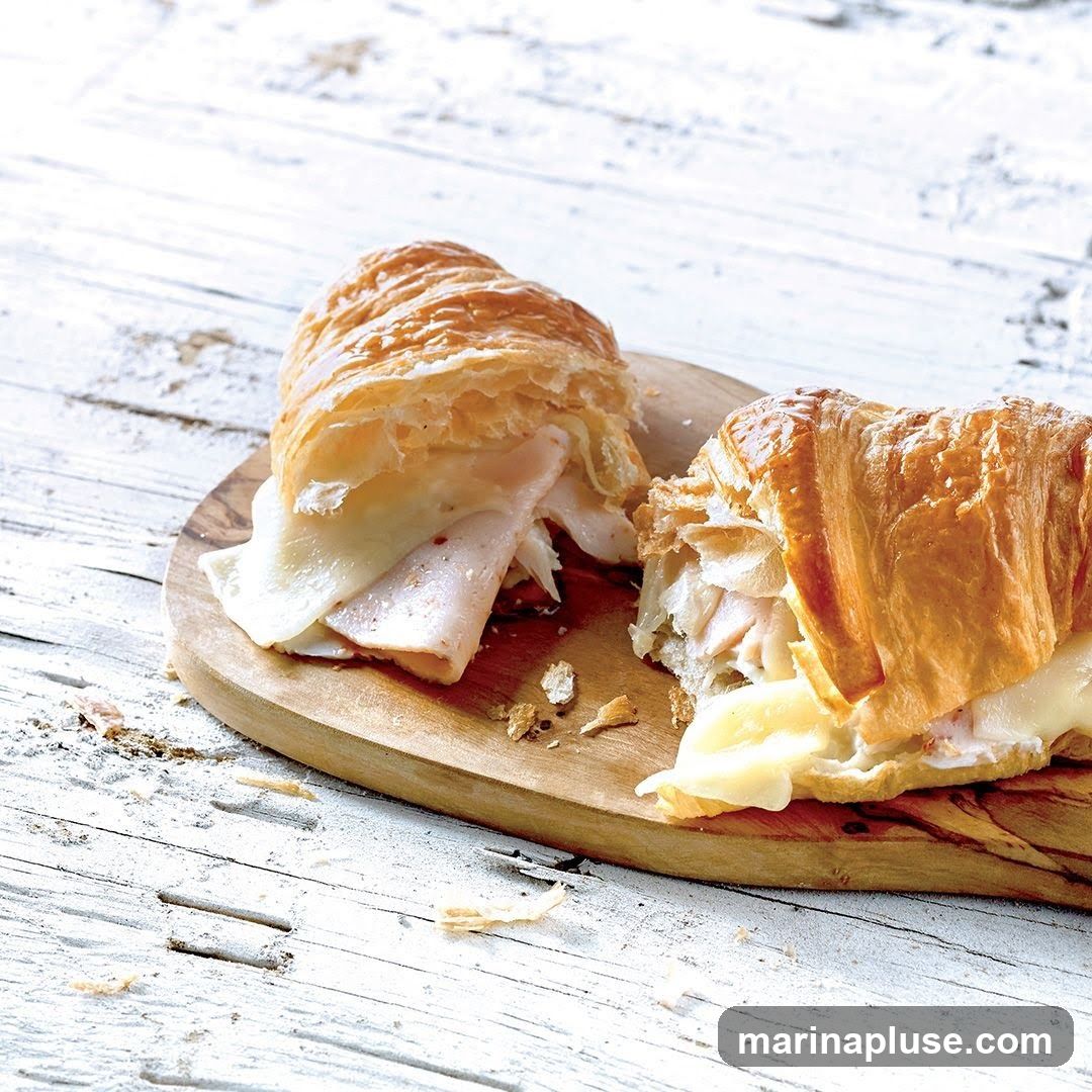 A croissant sandwich filled with turkey and cheese, cut in half and served on a wooden board. The background is a distressed white wooden surface.