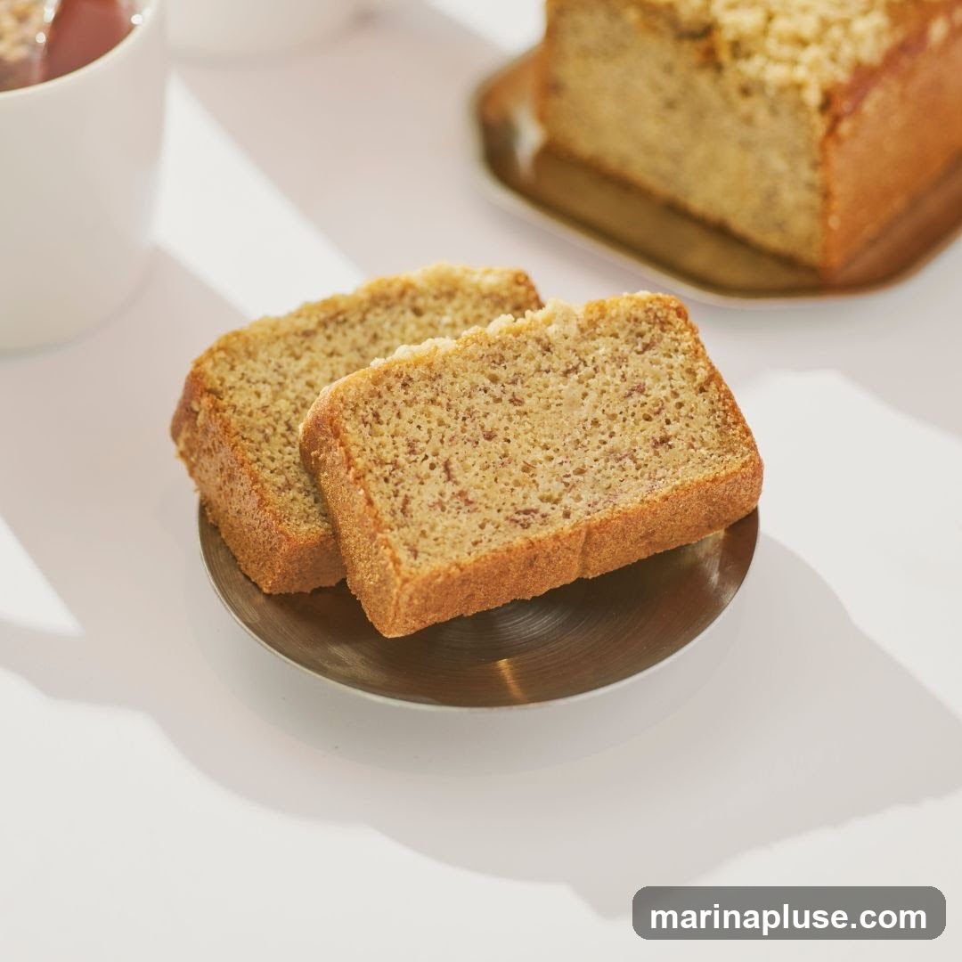 Two slices of banana bread are presented on a small, round, metallic plate. A larger loaf of banana bread, topped with a crumbly streusel, is visible in the background on a rectangular metallic tray. A white mug, partially filled with a reddish beverage, is in the upper left corner. The scene is set against a bright white surface, with soft shadows cast by the baked goods and mug, suggesting a well-lit, possibly indoor setting.