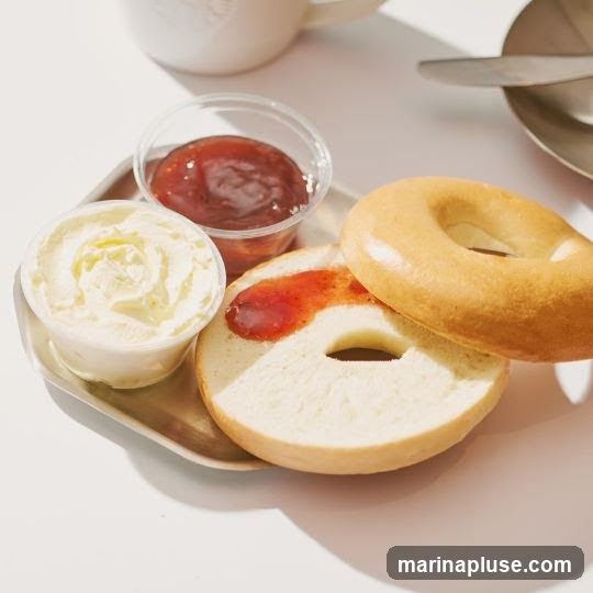A close-up shot of a plain bagel, sliced in half, with a dollop of cream cheese and a small container of jam. The bagel is spread with jam, and a second whole bagel sits beside it. A white mug with a subtle logo is visible in the background, along with a plate and cutlery.