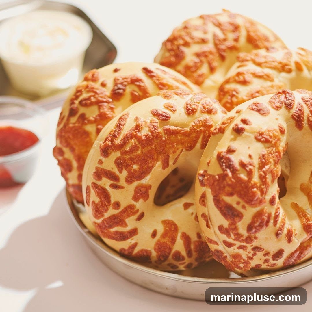 A close-up shot of several cheese bagels arranged on a silver plate. In the background, a small dish of jam and a container of cream cheese are visible. The bagels are golden brown with melted cheese baked onto their surface.