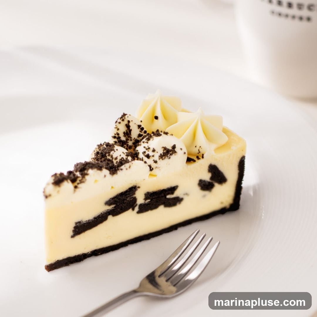 A slice of Oreo cheesecake on a white plate with a fork. The cheesecake has layers of cream cheese filling and crushed Oreos, topped with whipped cream and more crushed Oreos. A white mug with a logo is visible in the background.
