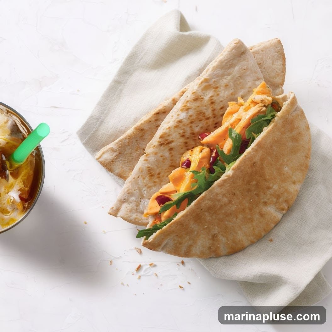 A close-up shot of a pita sandwich filled with orange filling, greens, and red pieces, served on a white cloth. To the left, a glass with a yellow drink and a green straw is visible. The background is a textured white surface.