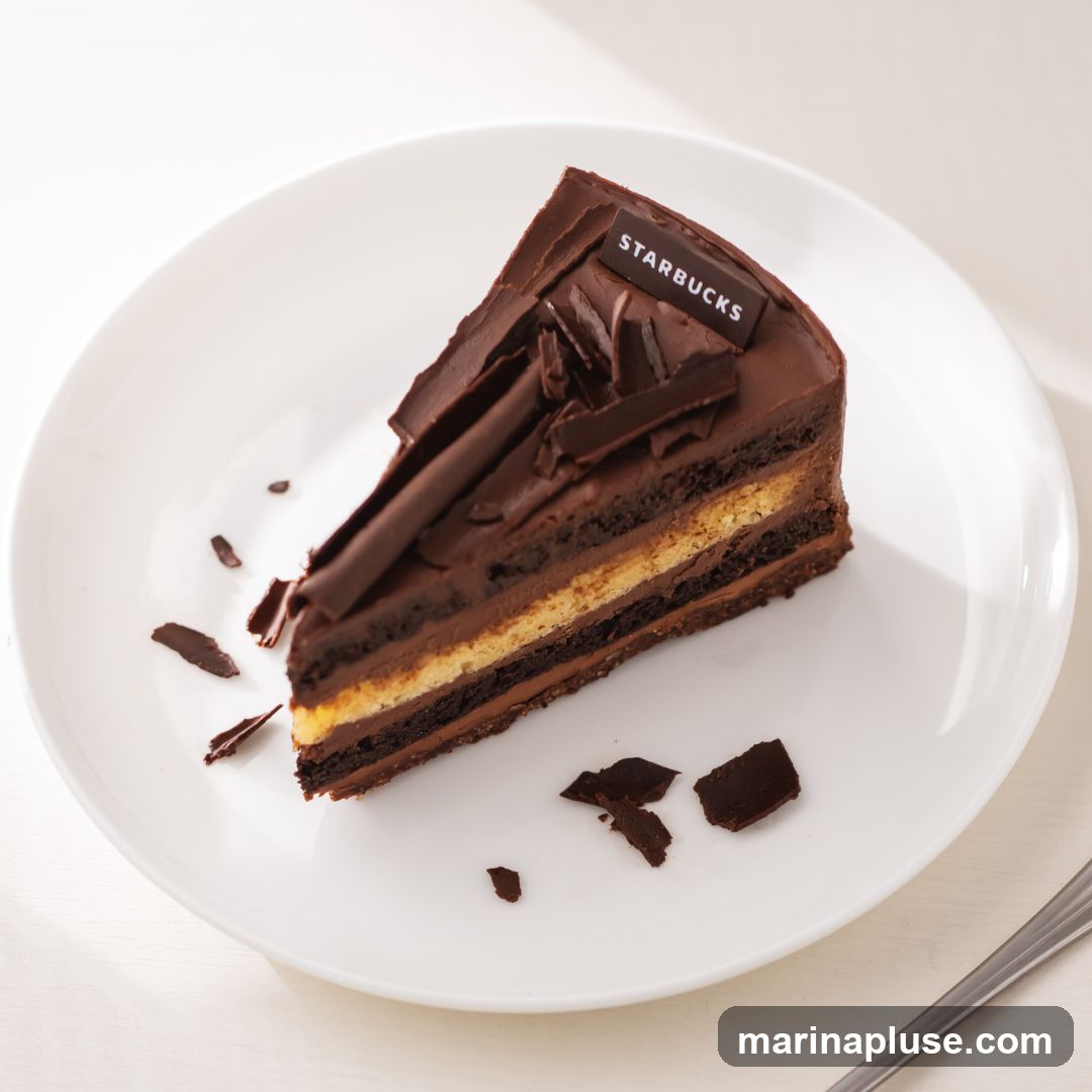 A slice of rich chocolate cake with layers of chocolate cake and cream, topped with chocolate shavings and a Starbucks branded chocolate plaque, served on a white plate.
