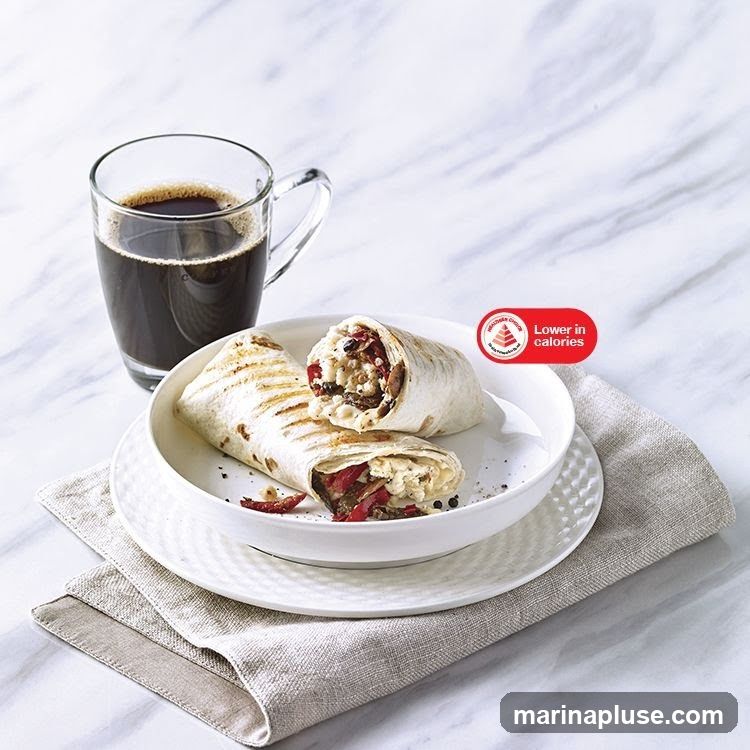Two grilled breakfast burritos are served in a white bowl on a white plate with a textured rim. A glass mug of black coffee sits to the left of the bowl. A linen napkin is placed under the plate. A red and white circular sticker with the text "Lower in calories" is visible on the right side of the image.