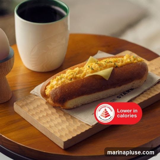 A close-up shot of a scrambled egg hot dog with a slice of cheese on top, served on a napkin on a wooden board. A white mug with a green rim and embossed logo is visible in the background, along with a portion of an egg in an egg cup. A red sticker on the napkin reads "Healthier Choice Lower in calories".