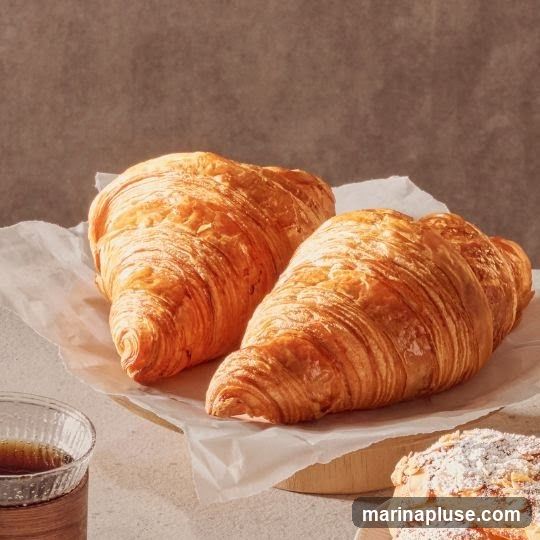 Two golden-brown croissants are displayed on white parchment paper, resting on a wooden stand. To the left, a glass of dark liquid, likely coffee, is visible. In the lower right corner, a pastry dusted with powdered sugar and almonds can be seen. The background is a textured, muted brown.
