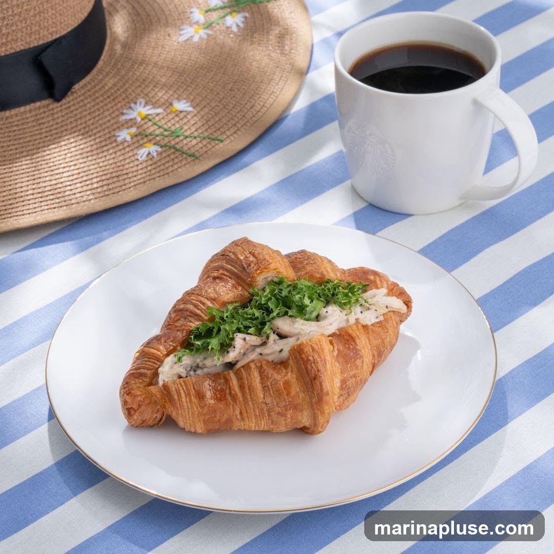 A close-up shot of a croissant sandwich filled with chicken salad and greens, served on a white plate with a gold rim. A white mug of black coffee is visible in the background, along with a straw hat adorned with daisies. The items are arranged on a blue and white striped tablecloth. The mug has a subtle Starbucks logo embossed on it.