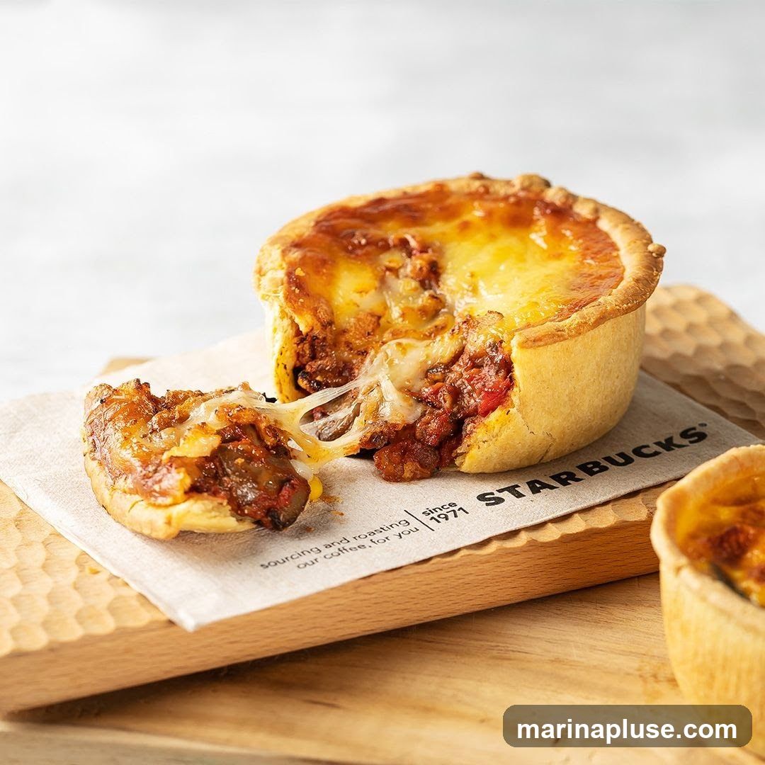 A close-up shot of a savory pie with a golden crust, filled with a rich meat and tomato mixture, topped with melted cheese. A piece of the pie has been removed, revealing the gooey cheese pull. The pie is served on a wooden board with a Starbucks branded napkin. Another small pie is visible in the background.