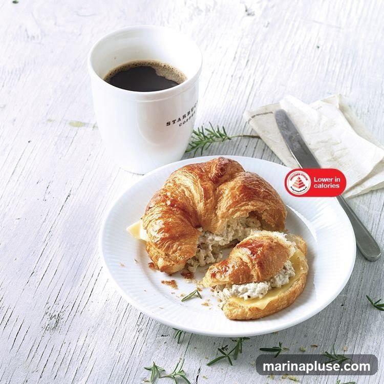 A croissant sandwich filled with a creamy mixture and cheese, served on a white plate with a sprig of rosemary. A white mug with "STARBUCKS COFFEE" visible is filled with black coffee. A napkin and knife are placed to the right of the plate. A red sticker indicates "Lower in calories".