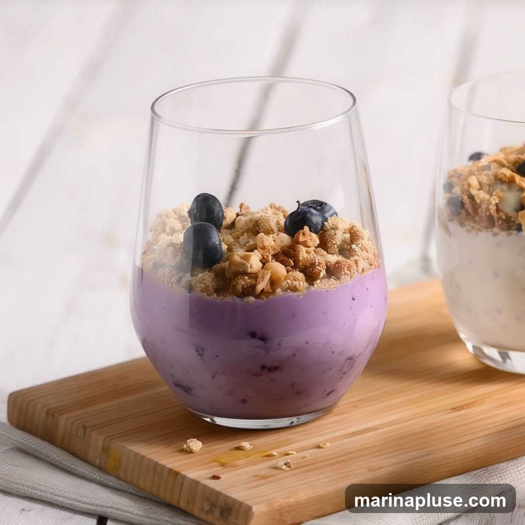 A close-up shot of a blueberry yogurt parfait in a clear glass, topped with granola and fresh blueberries. Another parfait is visible in the background. The glasses sit on a wooden board with a light-colored cloth underneath.