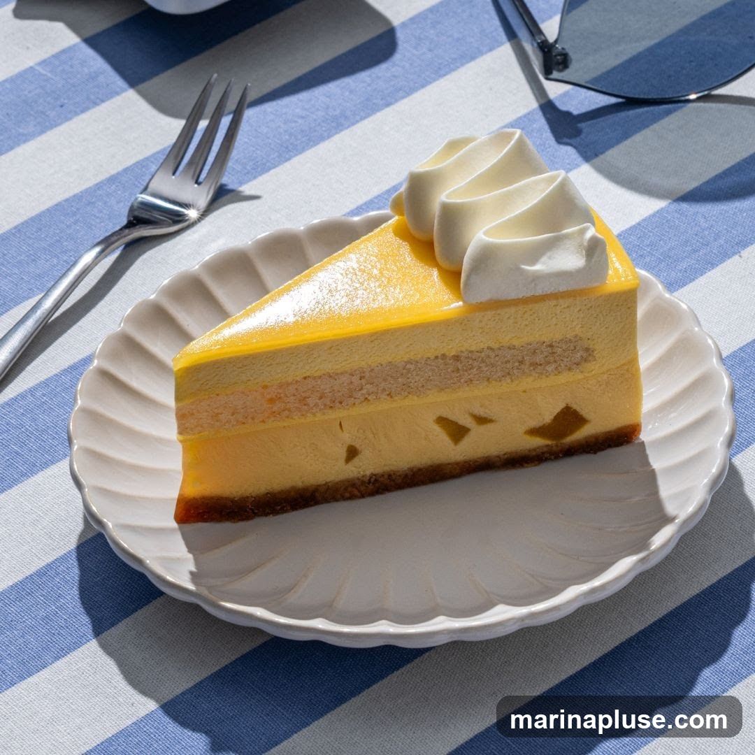 A slice of layered mango cake with whipped cream on top, served on a white scalloped plate. The cake has a yellow jelly layer, a sponge cake layer, and a creamy mango filling with visible mango chunks. It is placed on a blue and white striped tablecloth. A fork and sunglasses are also visible on the table.