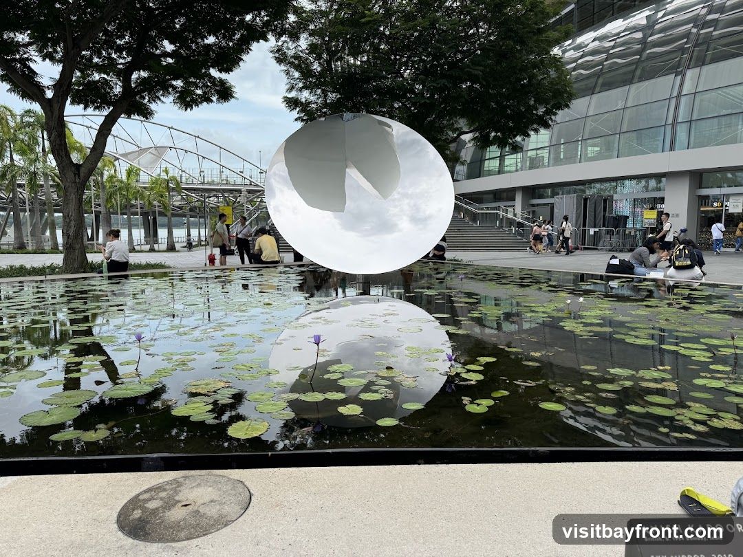 A modern outdoor scene featuring a reflective pool with lily pads and purple water lilies, mirroring a large metallic sphere sculpture. In the background, there is a contemporary glass building with people walking around, some sitting near the pool edge. The setting appears to be an urban park or plaza.