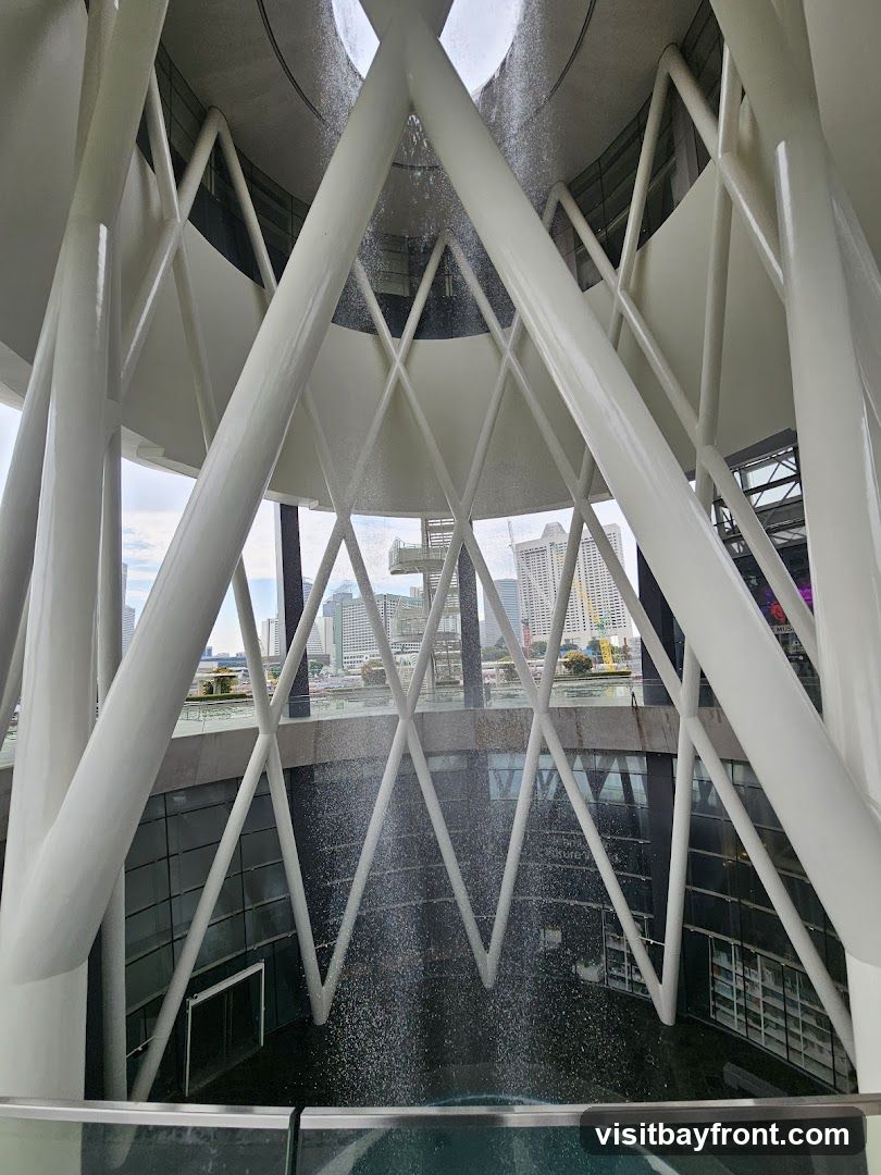 The image shows an interior space featuring a striking architectural design with white, angular beams forming a geometric pattern. A water feature cascades down the center, creating a dynamic visual element. Through the structure, a cityscape is visible in the background, suggesting an urban setting. The scene captures both the modern aesthetic and the surrounding environment.