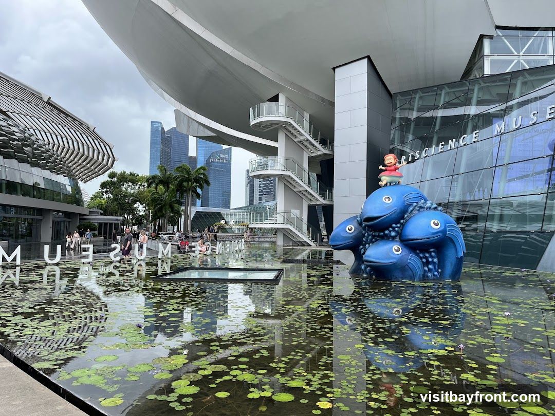The image shows the exterior of the Singapore Science Museum, featuring a large blue fish sculpture in a water feature covered with lily pads. The museum's modern architecture includes glass facades and curved structures. In the background, there are tall skyscrapers under an overcast sky. People can be seen walking near the entrance area.